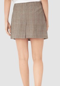 Mujer vistiendo una falda corta a cuadros marrón con un plisado en la parte trasera, de pie con los brazos relajados a los lados frente a un fondo sencillo.