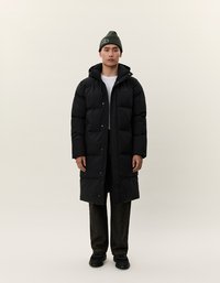 Giovane uomo che indossa un lungo cappotto imbottito nero, un berretto verde oliva, una camicia bianca, pantaloni scuri e scarpe nere, in piedi contro uno sfondo chiaro e uniforme.