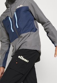 Szara i granatowa kurtka wiatrówka z niebieskimi suwakami, z kapturem i kontrastowymi panelami. Czarne spodnie z logo "ellesse" i kieszenią na zamek.
