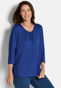 Blaues, langärmeliges Oberteil aus glattem Stoff mit Rundhalsausschnitt und Faltendetail vorne; kombiniert mit dunkelblauen Jeans.