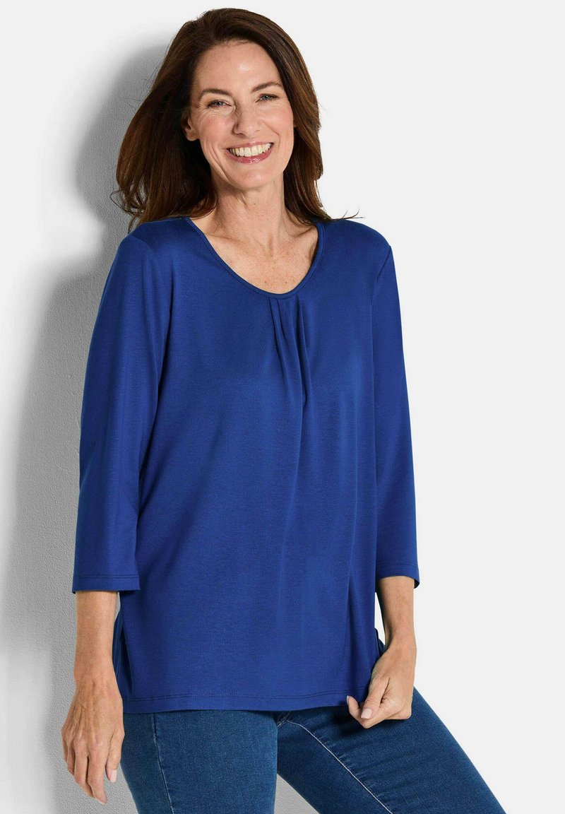 Blaues, langärmeliges Oberteil aus glattem Stoff mit Rundhalsausschnitt und Faltendetail vorne; kombiniert mit dunkelblauen Jeans.