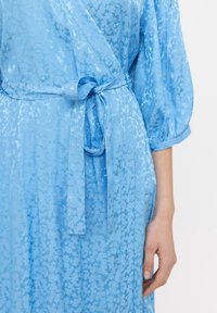 Jasnoniebieska sukienka kopertowa z teksturowanym, kwiatowym wzorem, z paskiem do zawiązania i bufkami. Wykonana z miękkiego, zwiewnego materiału.