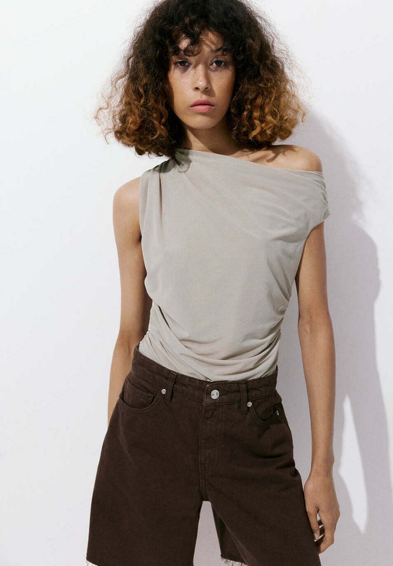 Femme aux cheveux bouclés portant un top gris clair une épaule et un short taille haute marron foncé, debout devant un fond blanc uni.