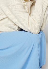Maglione lavorato a maglia color crema con costine testurizzate, abbinato a una gonna plissettata azzurro chiaro, con morbide pieghe e una finitura liscia.