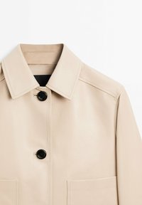 Beige læderjakke med krave, sort knaplukning og to forlommer. Glat tekstur og rene syninger.