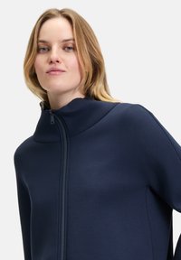 Marineblauer Reißverschlussjacke mit hohem Kragen, aus glattem Stoff gefertigt, mit minimalistischem Design und nahtlosen Schultern für einen modernen Look.