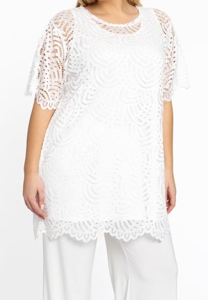 Personne portant une tunique en dentelle blanche avec des bords festonnés sur un pantalon blanc, montrant des motifs découpés complexes sur le tissu.