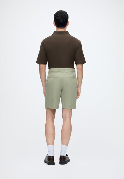 Homme de dos, portant une chemise à manches courtes marron foncé, un short vert clair, des chaussettes blanches et des chaussures marron foncé.