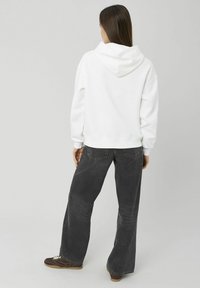 Femme aux longs cheveux bruns portant un sweat à capuche blanc, un jean noir ample et des baskets marron, debout de dos sur un fond uni.