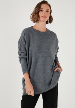 Oversized grijze gebreide trui met ronde hals, doorgezakte schouders en rafelige zoomdetails. Zachte textuur, lange mouwen met omgeslagen manchet.