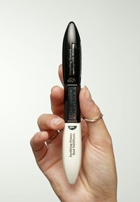 Mascarà noir à double embout avec une finition brillante. Une extrémité présente une section de primer blanche. Le texte détaillé sur l'emballage inclut les noms des produits.