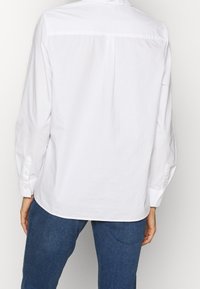 Weißes Hemd mit Knopfleiste, mit einem Rückenpasse, langen Ärmeln und Manschetten. Kombiniert mit blauen Jeans. Hat eine glatte Baumwolltextur.