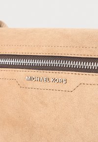 Tan semiš torba s srebrnim zadrgo in vtisnjenim logotipom "Michael Kors". Gladka tekstura z vidnimi šivi in podrobnimi poudarki.