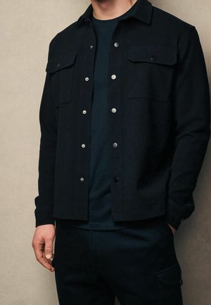 Mężczyzna ubrany w czarną, zapinaną na guziki kurtkę z klapami na kieszeniach na piersi, na czarnej koszuli, stojący na jednolitym beżowym tle.