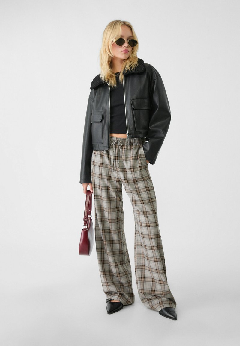 Blonde Frau, die Sonnenbrillen trägt, eine schwarze, kurz geschnittene Lederjacke, ein schwarzes Oberteil, karierte weite Hosen und spitze schwarze Schuhe anhat und eine rote Handtasche hält.