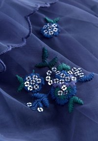 Tela azul marino transparente con diseño floral bordado en azul y verde, acentuado con lentejuelas plateadas circulares y borde festoneado.