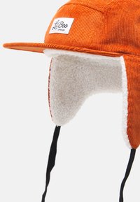 Orange corduroy cap with a flat brim and white, soft fleece ear flaps. Features black adjustable straps and a white logo patch on the front.