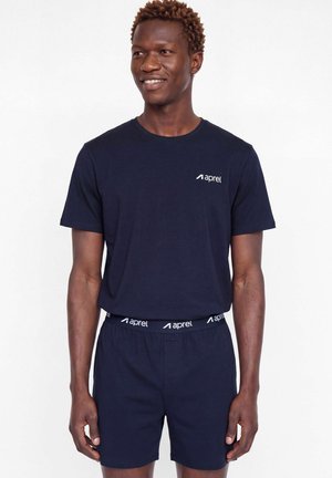 Un homme souriant porte un T-shirt bleu marine et un short assorti avec une ceinture élastique siglée, sur un fond blanc uni.