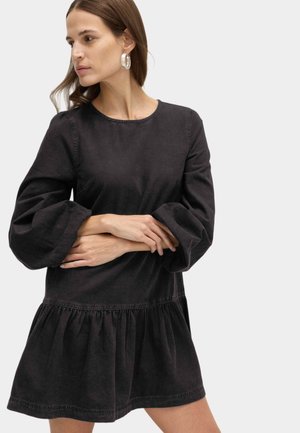 Femme aux cheveux châtain clair portant de grandes boucles d'oreilles argentées en forme d'anneaux et une robe noire à manches longues avec jupe froncée et manches bouffantes.