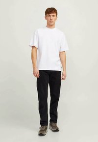 Joven de pie, vistiendo una camiseta blanca de manga corta, pantalones negros y zapatillas grises, contra un fondo claro y neutro.