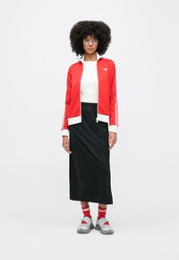 Sieviete ar cirtainiem matiem, valkā sarkanu sporta jaku, baltu topu, melnu garu svārku, sarkanas zeķes, pelēkas sandales un caurspīdīgas brilles, stāv uz balta fona.