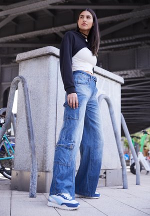 Jeans cargo a gamba larga in denim blu sbiadito con multiple tasche, abbinati a un crop top bianco e una felpa nera. Sneakers sportive completano il look.