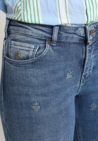 Blå denimjeans har en åtsittande design, framfickor och subtila broderade mönster på tyget. Texturen verkar något använd.