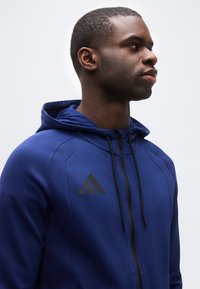 Νέος άνδρας με μπλε φούτερ με φερμουάρ και λογότυπο Adidas, κοιτάζοντας δεξιά, με φόντο απλό ανοιχτόχρωμο.