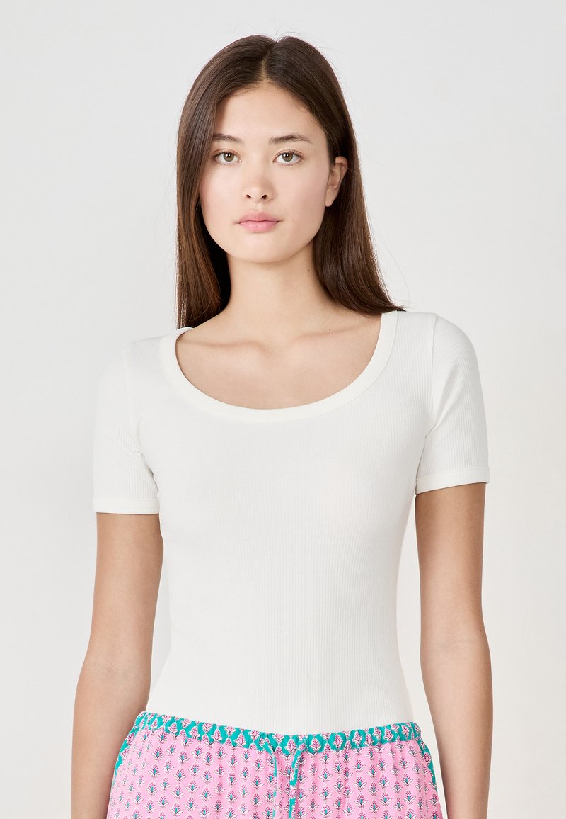 Top bianco a maniche corte con scollatura rotonda. Presenta un design attillato e una texture liscia. Abbinato a pantaloni a fantasia rosa e turchese.