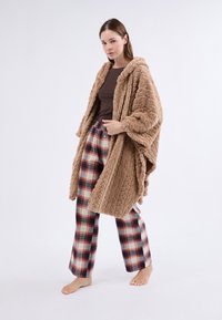 Hnědá kapucínová deka s texturovaným, nadýchaným designem. V kombinaci s kostkovanými kalhotami v červených a modrých tónech. Model stojí bosý na jednobarevném pozadí.