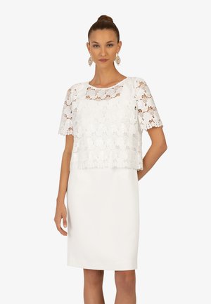 Weißes Spitzenoberteil mit kurzen Ärmeln über einem einfarbigen weißen Kleid. Die Spitze zeigt florale Muster und gewellte Kanten am Saum.