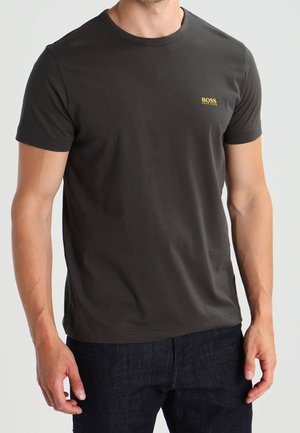 Mann trägt ein schlichtes dunkelgraues T-Shirt mit kurzen Ärmeln und kleinem gelbem "BOSS"-Logo auf der Brust, kombiniert mit dunkelblauen Jeans.