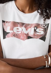 Osoba ve bílém tričku Levi's s květinovým logem a stříbrnými náramky na zkřížených rukou.