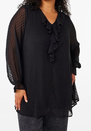 Black sheer blouse with dotted texture, ruffled V-neckline, and gathered cuffs worn over dark gray pants by a person.