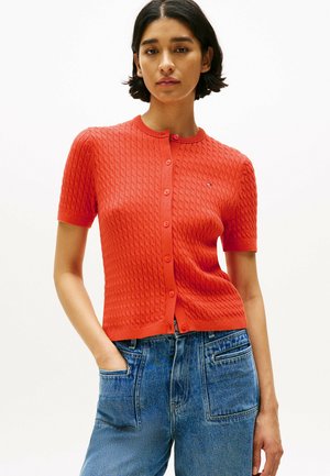 Cardigan rojo de manga corta con patrón de tejido texturizado, cierre de botones en la parte frontal y cuello redondo, combinado con vaqueros de talle alto azul.
