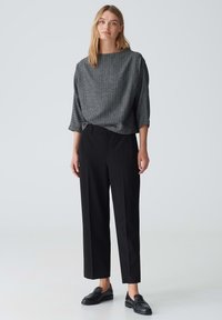 Grijze, gestructureerde top met brede mouwen, gecombineerd met hoge zwarte broek. Model in zwarte loafers staat tegen een lichte achtergrond.