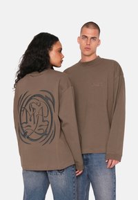 Langärmliges braunes Baumwoll-T-Shirt mit einem großen schwarzen Grafikdesign auf dem Rücken und einem kleinen gestickten Logo auf der Vorderseite.