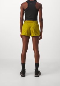 Krótkie, jasnożółte spodenki sportowe z elastycznym pasem, wykonane z lekkiego materiału, pokazane od tyłu obok ciemnych sportowych skarpet i butów.