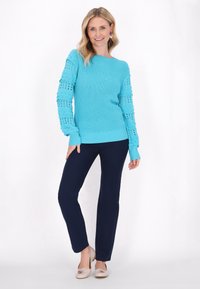 Pulover tricotat turcoaz cu mâneci texturate, asortat cu pantaloni bleumarin și încălțăminte de culoare deschisă. Hainele prezintă accente de tricotare deschisă.