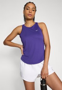 Lila idrottstopp med rund hals och ärmlös design. I kombination med vita shorts, håller en svart vattenflaska. Nike-logotyp synlig.