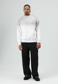 Witte trui met een gradiënt grijs patroon en kleine zwarte versieringen. Gecombineerd met losse zwarte spijkerbroek en dikke zwarte schoenen.