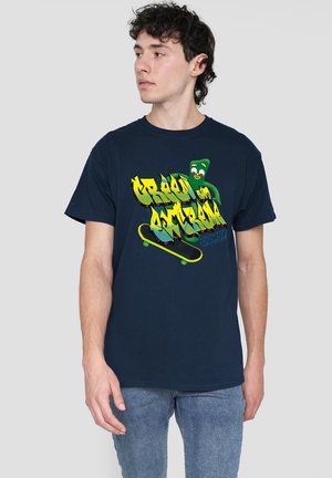 Junger Mann mit dunklem, lockigem Haar, der ein navyblaues T-Shirt trägt, das mit buntem "Green on Scrum" Graffiti und einem grünen Cartoon-Charakter auf einem Skateboard bedruckt ist.