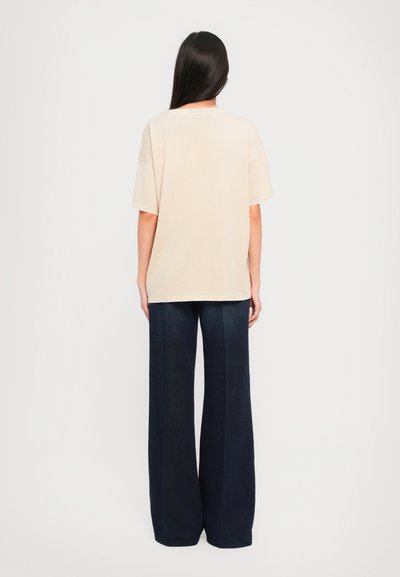 Camiseta oversized beige de manga corta y escote redondeado, combinada con pantalones vaqueros de pierna ancha en azul oscuro. Textura de tela suave.