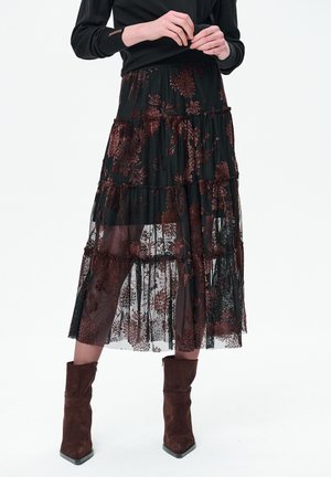 Kobieta ubrana w czarną bluzkę z długim rękawem, prześwitującą czarną spódnicę midi z ciemnym kwiatowym wzorem oraz brązowe zamszowe botki za kostkę.