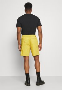 Žluté kraťasy s elastickým pasem, bočními kapsami a jednoduchým designem. Nosí se s černým tričkem a černými botami.