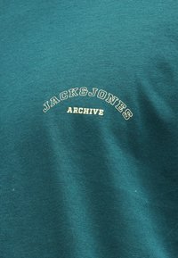 Τυρκουάζ ύφασμα με κρεμ χρωματιστό κείμενο "JACK & JONES" που είναι τοξοειδώς τοποθετημένο πάνω από το "ARCHIVE" σε κεφαλαία γράμματα, κεντραρισμένο στην πάνω περιοχή του στήθους.
