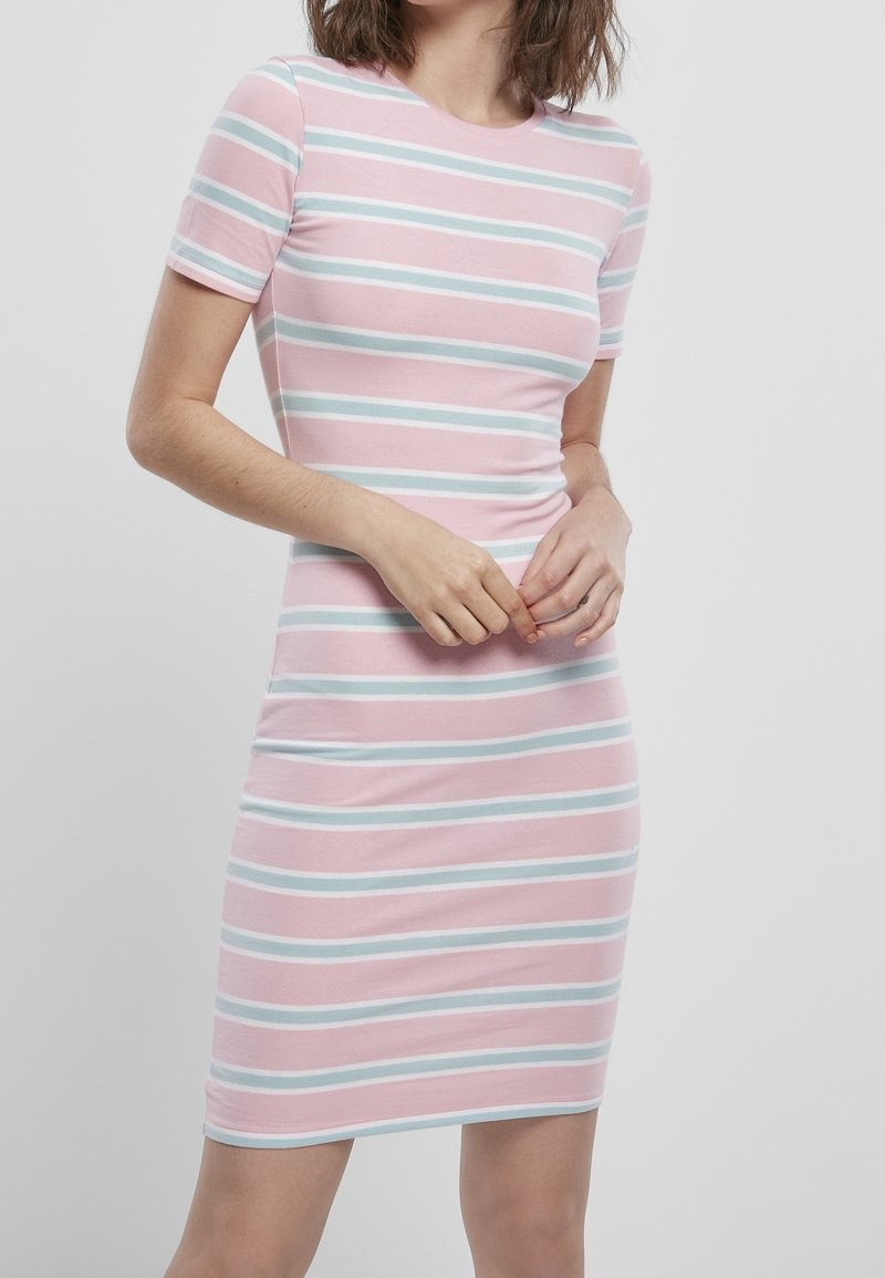 Femme portant une robe rose avec des rayures horizontales blanches et bleu clair, debout, les mains légèrement jointes devant elle.