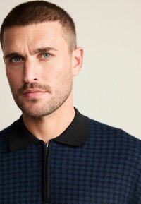 Pánska polokošeľa na zips s motívom psieho zubov v námorníckej modrej a čiernej farbe s čiernym golierom. Detailný záber na tvár modela s krátkymi vlasmi a bradou.