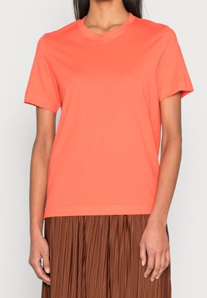 Mujer vistiendo una camiseta coral de manga corta y cuello redondo, combinada con una falda marrón plisada, mostrada desde el cuello hasta la cintura.