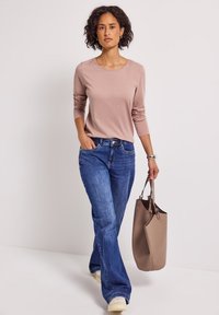 Femme aux cheveux bouclés marchant avec une chemise à manches longues rose pâle, un jean bleu, des chaussures blanches et portant un sac à main taupe d'une main.